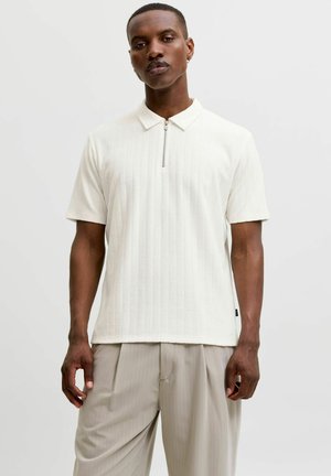 Homme portant une chemise blanche côtelée à manches courtes avec col à fermeture éclair et un pantalon beige clair à fines rayures, debout devant un fond uni.
