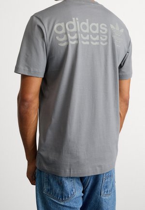 Mężczyzna ubrany w szary t-shirt Adidas z odblaskowym logo i koniczynką na górnej części pleców, w połączeniu z niebieskimi dżinsami.