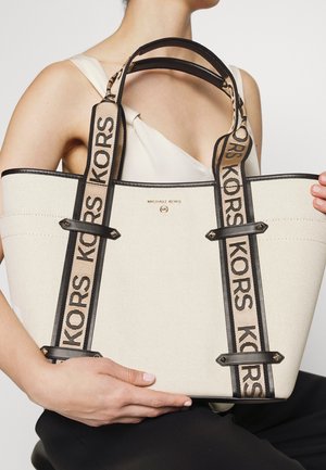 Femme tenant un grand sac cabas beige Michael Kors avec des bordures noires et de larges anses d'épaule à logo.