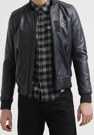 Veste bomber en cuir noir avec des poignets et un col côtelés, fermeture éclair à l'avant, poche sur la poitrine, portée par-dessus une chemise à carreaux noire et grise.