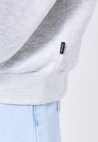 Sudadera gris con un acabado texturizado, que presenta una etiqueta de marca negra. Combinada con jeans de mezclilla azul claro, que muestran costuras y un ligero deshilachado.