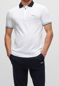 Chemise polo blanche avec col marine et accents bleu clair sur les manches. Logo sur la poitrine et portée avec un pantalon sombre. Matière en coton lisse et ajustée.