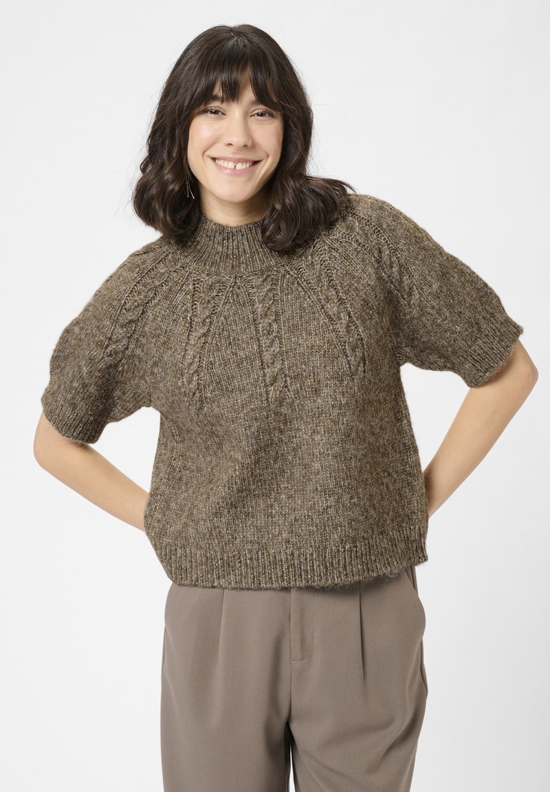 Smilende kvinde med skulderlangt mørkt hår iført en brun kortærmet strikket sweater og taupe højtaljede bukser, hænderne bag ryggen.