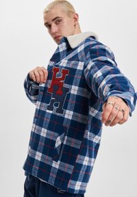 Blå og hvidternet flannelskjorte med en beige sherpa-krave, prydet med røde og marineblå broderede bogstaver på forsiden og en sidelomme.