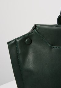 Ciemnozielona skórzana torba z narożnikami i przeszytymi brzegami oraz pasującym okrągłym guzikiem na jasnym, jednolitym tle.