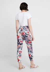 Un pantalon de jogging à motif floral présente une base blanche avec des fleurs tropicales rouges et bleues, des poignets élastiques et une coupe décontractée, associé à un t-shirt blanc uni.