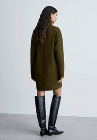 Vestito a maglia oversize verde oliva con maniche lunghe, tessuto strutturato e collo alto, abbinato a stivali neri in pelle al ginocchio.