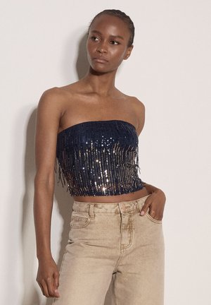 Kobieta z plecionymi włosami, ubrana w bezramkowy granatowy top cekinowy i beżowe spodnie z wysokim stanem, stojąca z jedną ręką w kieszeni.