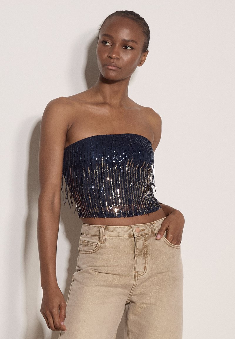 Femme aux cheveux tressés portant un top bustier bleu marine à sequins et un pantalon beige taille haute, debout avec une main dans la poche.