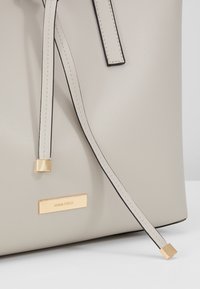 Beige håndtaske med struktureret materiale, der har sorte accenter, et guldsymbol med teksten "ANNA FIELD" og en lang rem med firkantet ende.