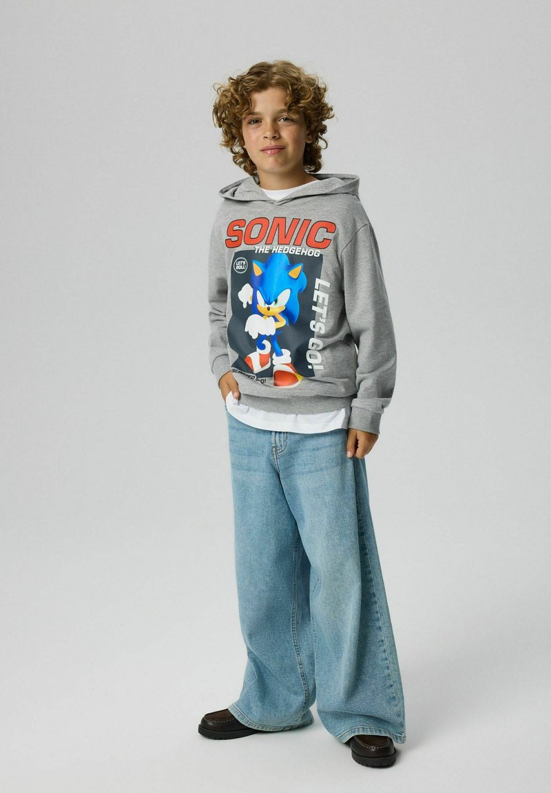 Garçon aux cheveux bouclés portant un sweat à capuche gris Sonic the Hedgehog et un jean large bleu clair, debout devant un fond uni.