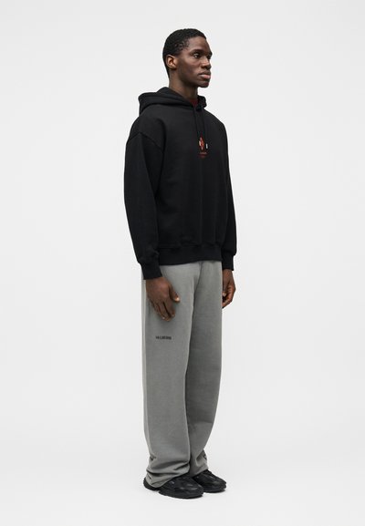 Juodas su gobtuvu džemperis su priekyje esančiu piešiniu, derinamas su laisvomis pilkomis sportinėmis kelnėmis su logotipu. Modelis dėvi juodus sportinius batelius.