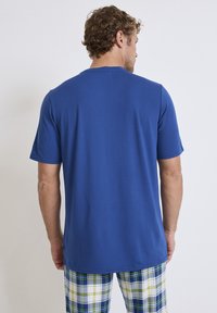 T-shirt blu a maniche corte realizzato in tessuto morbido, con collo rotondo. Il modello indossa pantaloni da pigiama a quadri nei colori blu, verde e bianco.