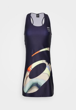 Nainen, jolla on päällään hihaton tumma urheilumekko abstraktilla kuviolla ja valkoiset lenkkarit, seisoo kädet selän takana harmaalla lattialla.