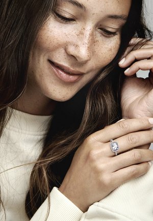Anello con diamante a forma quadrata circondato da un alone di pietre più piccole, montato su una delicata fascia. La mano è appoggiata su un leggero maglione, con lunghi capelli parzialmente visibili.