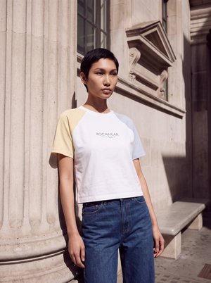 Une femme porte un T-shirt blanc aux manches jaunes et bleu pâle et un jean bleu, près d’un bâtiment en pierre baigné de soleil.