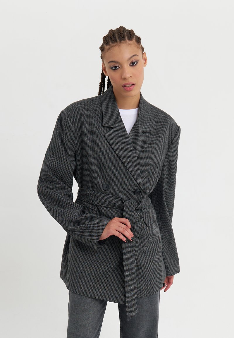 Grå rutete blazer med belte i midjen, dobbeltradet front, brede revers og lange ermer. Har et teksturert stoff.