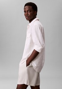 Junger Mann in einem weißen karierte Langarmhemd mit hochgekrempelten Ärmeln und cremefarbigen Shorts, steht mit den Händen in den Taschen vor einem grauen Hintergrund.