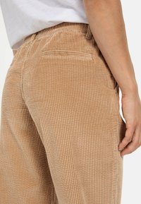 Sennepsbrune kordfløjlbukser med lodret ribbestruktur. Har lommer og elastisk talje, med en glat overflade på stoffet.