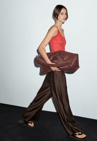 Red ribbed tank top, brown wide-leg trousers, holding a textured dark red bag. Walking on a dark floor with a light background.
