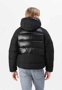 Doudoune noire avec une texture brillante et matelassée, dotée d'une capuche et d'un accent logo sur le col, associée à un jean en denim clair.