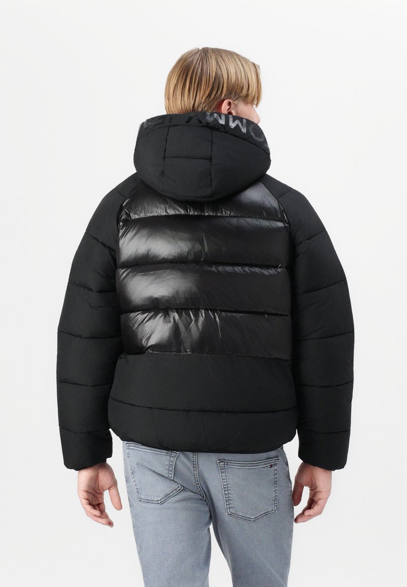 Schwarze Pufferjacke mit glanzvollem, gestepptem Textil, ausgestattet mit Kapuze und Logo-Detail am Kragen, kombiniert mit hellen Denim-Jeans.