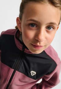 Fleecevest in roze en zwart, met een ritssluiting aan de voorkant, een hoge kraag en een logodetail op de borst. Zachte textuur en sportief ontwerp.