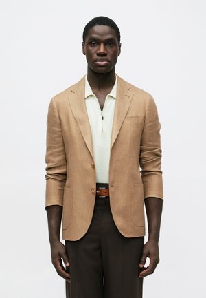 Muž v béžovom saku cez svetlo krémovú zipovú košeľu, tmavohnedé nohavice a zodpovedajúci opasok, stojaci pred jednoduchým bielym pozadím.