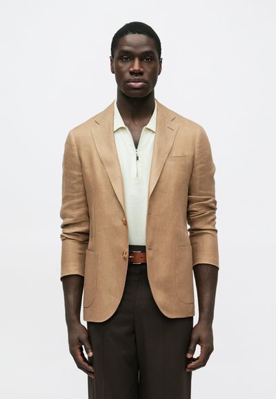 Homme portant un blazer beige par-dessus une chemise zippée crème clair, un pantalon marron foncé et une ceinture assortie, debout devant un fond blanc uni.