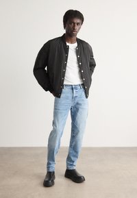 Chaqueta bomber negra con botones de presión plateados, camiseta blanca, jeans de mezclilla azul claro y zapatos negros sin cordones. Textura suave, ajuste relajado.