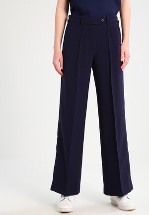 Granatowe spodnie o szerokich nogawkach wykonane z gładkiej tkaniny, z zapięciem na guzik, plisami i bocznymi kieszeniami. Połączone z białymi trampkami.