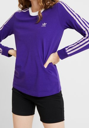 Osoba ubrana w fioletową koszulkę Adidas z długim rękawem z jasnoróżowymi paskami na rękawach i czarne szorty, ręce spoczywają na biodrach.