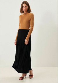 Brun ribbet langærmet top kombineret med en sort plisseret maxiskirt. Modellen har bordeauxfarvede hæle sandals på. Minimalistisk lys baggrund.