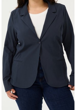 Kvinde iført en figursyet navyblå blazer, knappet én gang over en hvid top og mørkeblå jeans, stående med hænderne nær hofterne.