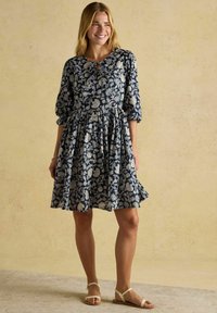 Frau lächelt in einem blumigen blauen Kleid mit Puffärmeln und steht auf einem hellen Boden vor einem sanft strukturierten Hintergrund.