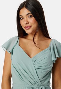 Licht teal wikkeljurk met korte vlinder mouwen, een V-halslijn en geplooide details bij de taille voor een getailleerd silhouet.