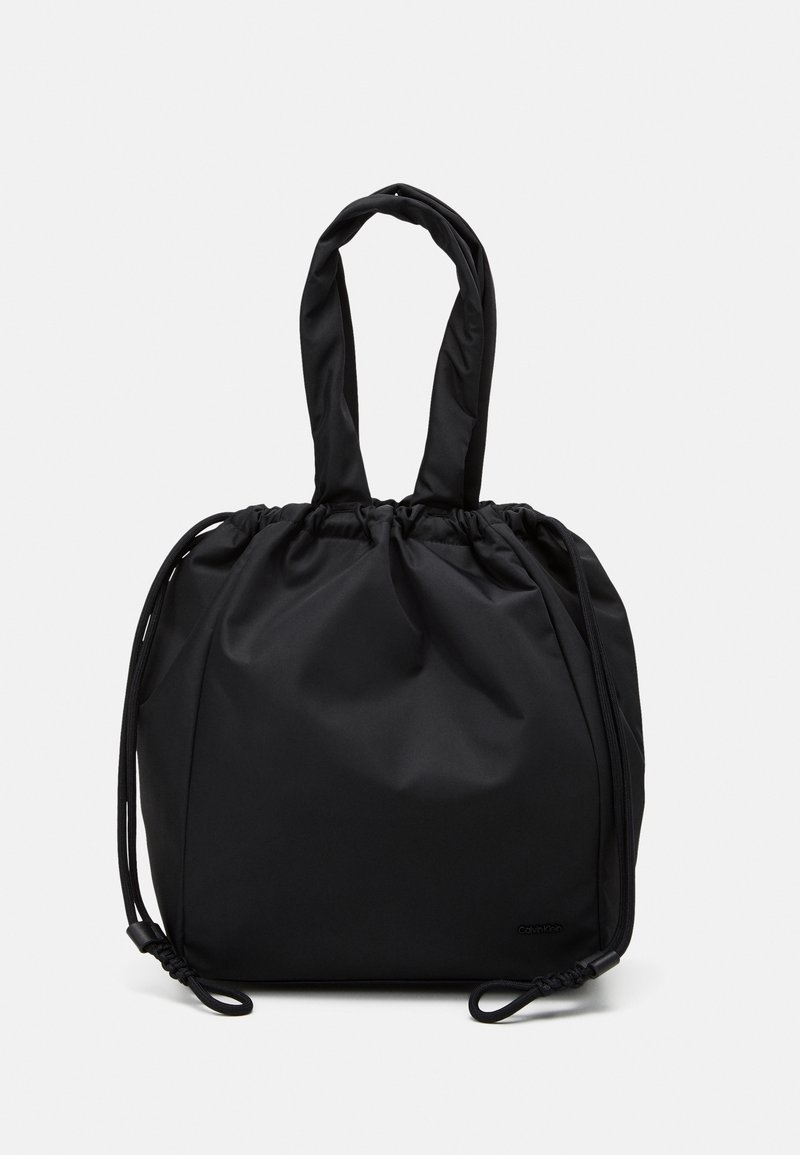 Sac fourre-tout en tissu noir doux avec haut froncé, double anse d'épaule et côtés à cordon réglable sur un fond clair uni.