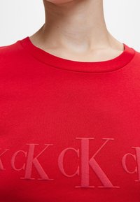 Camiseta de algodón roja con cuello redondo, con letras en relieve de un tono de rojo ligeramente más oscuro en el pecho. Textura suave.