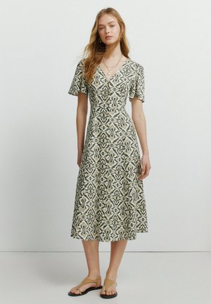 Kvinde iført grøn og hvid mønstret midikjole med korte ærmer og tanfarvede sandaler, der står mod en hvid baggrund.