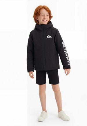 Дете с червена коса, облечено в черно качулково яке Quiksilver, черни къси панталони и бели маратонки, стои и се усмихва, гледайки настрани.