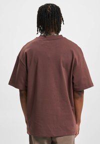 Camiseta de algodón marrón con un corte relajado, mangas cortas y cuello redondo. Presenta una textura suave y detalles de costura mínimos.