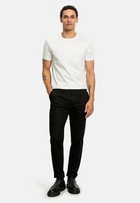 Weißes T-Shirt mit kurzen Ärmeln, kombiniert mit schwarzen Hosen und schwarzen Schuhen. Der Stoff wirkt glatt und die Passform ist maßgeschneidert.