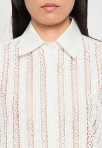 Moteris vilkinti baltą nėrinių bluzoną su vertikaliomis gėlių raštais ir tvirtu smailiu apykakle, kurios priekyje užsegta.