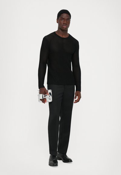 Άνδρας που φοράει μαύρο sheer μπλουζάκι μακρύ μανίκι και μαύρο παντελόνι, κρατώντας μια μικρή ανακλαστική ασημένια τσάντα, στέκεται μπροστά σε ένα απλό λευκό φόντο.