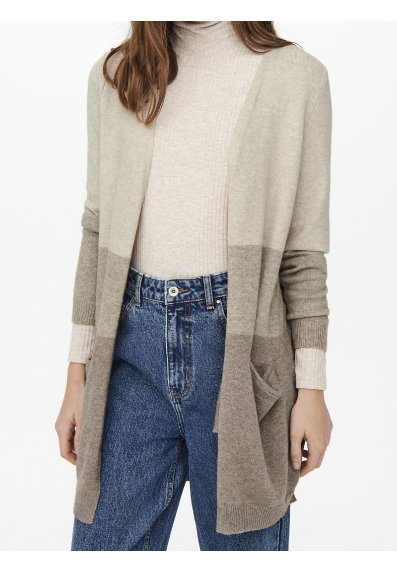 Femme portant un cardigan beige et marron à blocs de couleurs sur un col roulé beige clair et un jean bleu taille haute, debout sur fond blanc.
