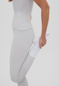 Top y leggings ajustados de color gris claro con una textura suave. Una mano agarra un calcetín blanco en el pie, enfatizando una pose de estiramiento.