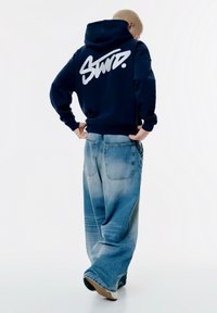 Felpa blu navy con logo bianco sulla schiena, abbinata a jeans chiari a gamba larga. La felpa ha un cordino e polsini a costine.