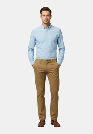 Man staat met handen in de zakken, draagt een lichtblauw overhemd met knopen, beige broek en bruine schoenen tegen een effen achtergrond.
