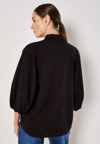 Schwarze Bluse mit hohem Hals, puffärmel und lockerer Passform. Glattes Material mit subtilen Nähdetails.