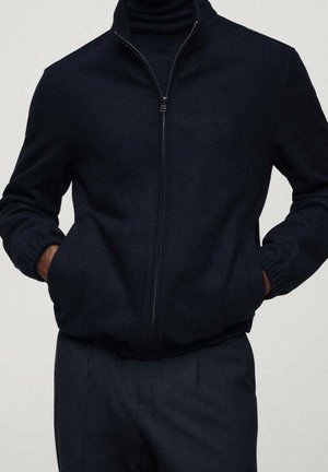 Homme portant une veste noire à fermeture éclair avec des poignets élastiques et les mains dans les poches, associée à un pantalon foncé.